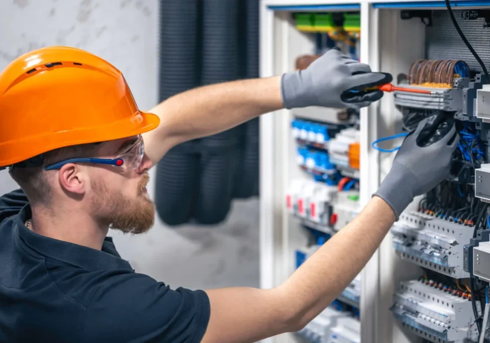 Electrician fixing wiring inside an electrical panel while wearing safety gear.